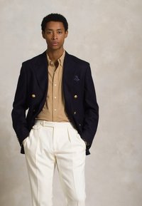 Blazer azul marinho de duas fileiras com botões dourados, camisa bege por baixo e calças cream plissadas. Lenço de bolso com padrão de poás.