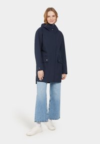 Marineblauer wasserdichter Mantel mit einem Reißverschluss vorne, zwei großen Fronttaschen und Kapuze. Kombiniert mit hellblauen Schlagjeans und weißen Sneakern.