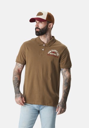 Homme barbu avec des tatouages, portant un polo marron Von Dutch et une casquette trucker marron et beige, regardant sur le côté.