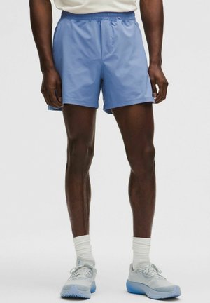 Mann trägt hellblaue Sportshorts, weiße Kniestrümpfe und weiße Sportschuhe mit blauen Sohlen, steht vor einem einfarbigen Hintergrund.