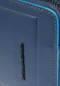 Portafoglio in pelle blu dalla texture liscia, con logo nero in rilievo e cuciture blu a contrasto lungo i bordi. Chiusura con zip visibile.