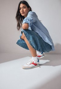 Fehér sportcipő piros és kék részletekkel, bézs talppal, fűzőkkel és texturált anyagból; kék-fehér csíkos inggel és farmer szoknyával párosítva.