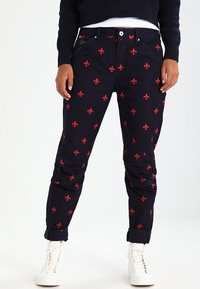 Černé kalhoty s červeným vzorem fleur-de-lis, s klasickým střihem, pěti kapsami a ohrnutými lemy, v kombinaci s bílými teniskami.