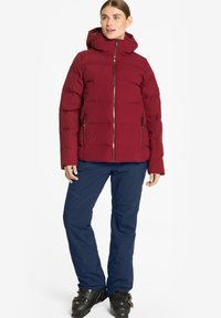 Doudoune rouge avec capuche, fermeture éclair à l'avant et poches latérales ; associée à un pantalon bleu marine et des bottes de ski noires. Texture lisse, design isolé.