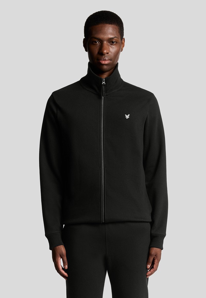Zwarte zip-up jas gemaakt van zacht materiaal, met een hoge kraag en een klein geborduurd logo op de linkerkant van de borst.