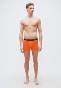 Νεαρός άνδρας που στέκεται ξυπόλητος, φορά φωτεινά πορτοκαλί εσώρουχα τύπου μποξέρ με μαύρη ζώνη μέσης που φέρει την επιγραφή «Polo Ralph Lauren».