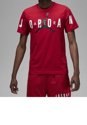 Mężczyzna w czerwonym t-shircie Jordan i dopasowanych szortach z czarnym logo Jumpman i białym napisem z przodu.