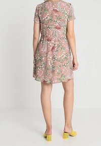 Vestido rosa de manga corta con patrón floral verde, cintura entallada y falda acampanada. Cierre de botón en la parte posterior y textura suave. Llevado con tacones amarillos.