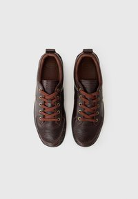Sneaker in pelle marrone con superficie texturizzata, dotate di lacci marroni, occhielli dorati e un logo sulla linguetta. Design della punta liscia e arrotondata.