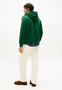 Joven de pie de espaldas, vistiendo un hoodie verde, pantalones blancos, camisa de cuadros y zapatos marrones, sobre un fondo blanco.