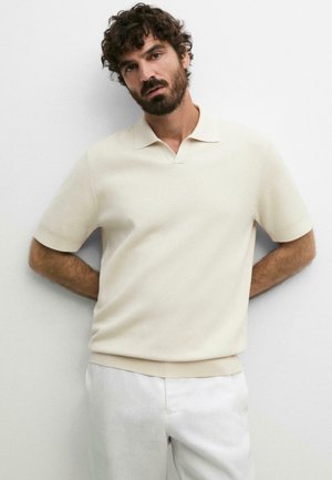 Uomo con capelli ricci e barba che indossa una polo beige a maniche corte e pantaloni bianchi, in piedi contro un muro bianco semplice.