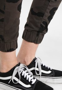 Kamouflage joggers i mörkgrön och svart tyg med elastiska muddar, tillsammans med svarta sneakers med vita detaljer och snörning.