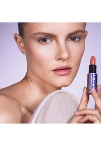 Un rouge à lèvres dans un tube métallique violet avec un capuchon noir, présentant une teinte nude. Le modèle a la peau claire et porte un maquillage léger pour les yeux.