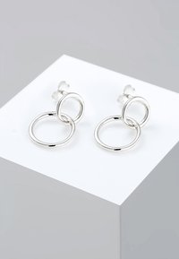 Pendientes de metal plateado que presentan dos aros circulares interconectados, acabado pulido y cierre de apoyo. Textura suave con bordes redondeados.