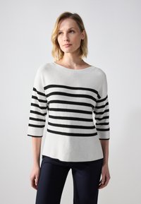 Pull en tricot blanc avec des rayures noires horizontales, encolure bateau et manches trois-quarts, associé à un pantalon sombre.