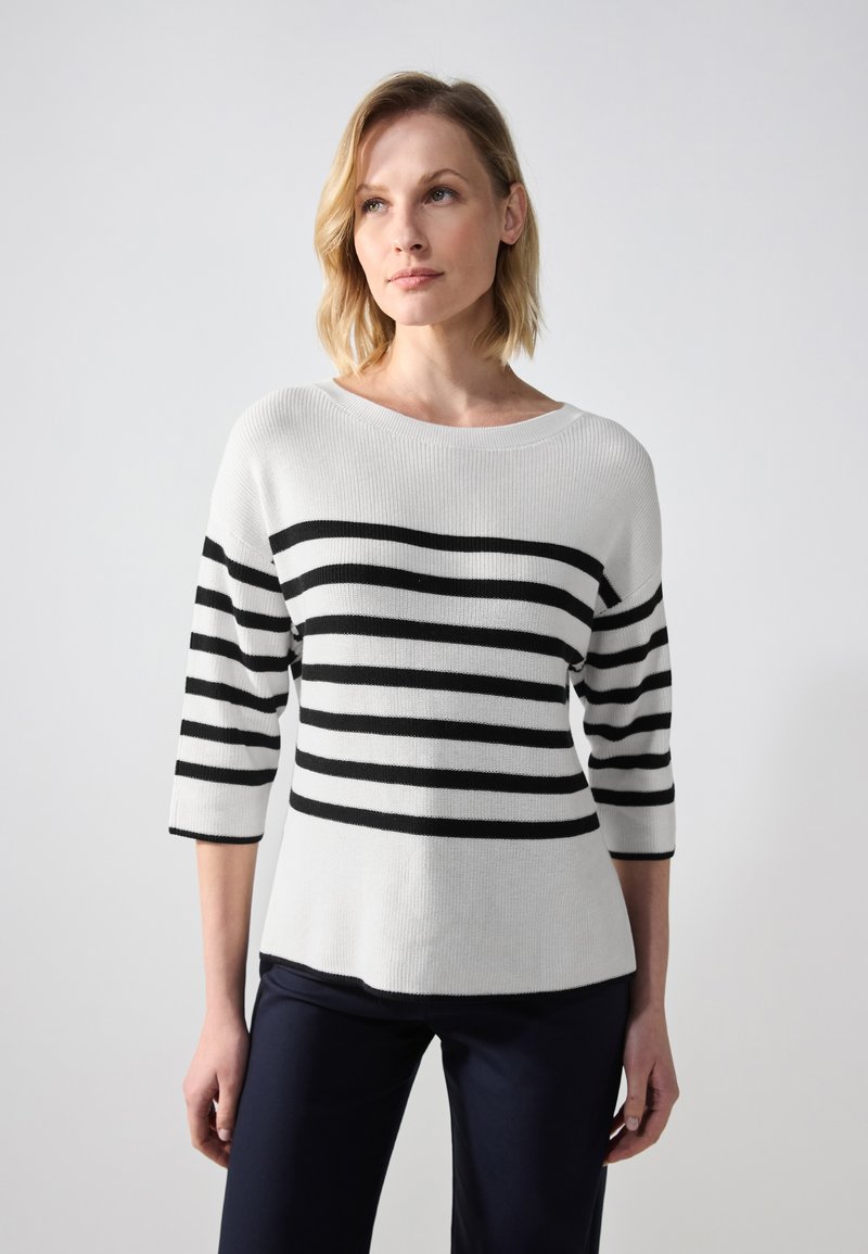 Pull en tricot blanc avec des rayures noires horizontales, encolure bateau et manches trois-quarts, associé à un pantalon sombre.