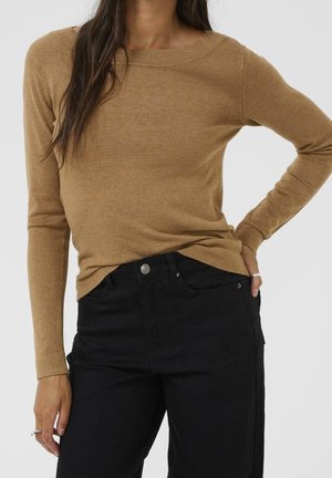 Femme portant un haut camel marron à manches longues ajusté, rentré dans un jean noir taille haute, main reposant dans la poche, sur un fond uni.