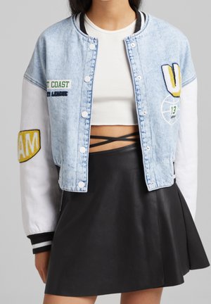 Veste bomber en denim avec un corps bleu clair, des manches blanches et des patchs brodés. Associée à une jupe en faux cuir noire.