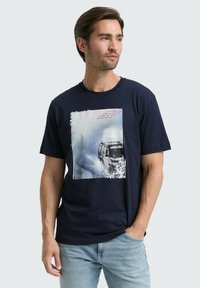 Tricou din bumbac de culoare bleumarin, cu un imprimeu grafic pătrat reprezentând un vehicul într-un peisaj acoperit de zăpadă, având margini uzate și accente de text.
