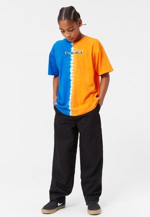 Koszulka z krótkim rękawem w niebiesko-pomarańczowy wzór tie-dye z białymi akcentami, zestawiona z czarnymi luźnymi spodniami i czarnymi butami.
