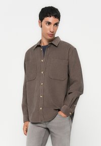Chemise en velours côtelé marron avec deux poches poitrine, devant à boutons et manches longues ; texture verticale légèrement côtelée ; boutons de couleur contrastante.