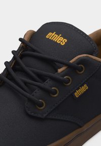Close-up of black Etnies sneaker with brown trim, black laces, metal eyelets, and yellow branding on tongue and side tag.