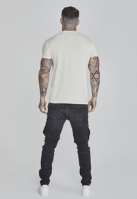 Tricou alb cu mânecă scurtă combinat cu pantaloni cargo negri, având mai multe buzunare, purtându-se cu adidași albi. Fundalul este simplu.
