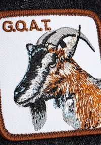 Patch brodé représentant une tête de chèvre en fils marron et noir sur un fond blanc, avec "G.O.A.T." au-dessus en marron.