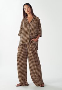 Brązowa, wzorzysta, oversize'owa koszula oraz dopasowane szerokie spodnie z geometrycznym wzorem, wykonane z lekkiej tkaniny. Brak widocznych elementów metalowych.