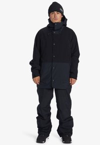 Donkerblauwe sneeuwjas met knopen voorkant, grote zakken en capuchon, gecombineerd met loszittende donkere broeken en zwarte laarzen.