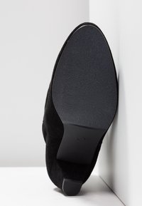 Suela de zapato de ante negra encarada hacia adelante, reposando contra una pared blanca en una esquina, mostrando suela de textura suave y pequeño tacón.