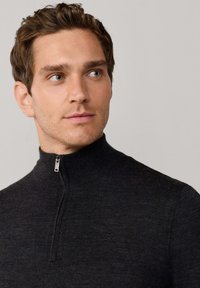 Donkergrijze ritsvest met hoge kraag, gemaakt van gestructureerde stof, met een subtiele logo op de borst. De rits heeft metalen hardware.