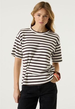 Frau mit langen blonden Haaren trägt ein schwarz-weiß gestreiftes T-Shirt, schwarze Hose, goldene Ohrringe und mehrere auffällige Armbänder am Handgelenk.