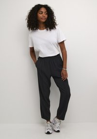 T-shirt en coton blanc associée à un pantalon élastique gris foncé et des baskets grises. Cheveux bouclés et main reposant sur la poche du pantalon.