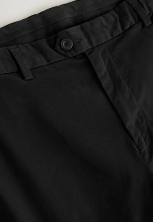 Juoda kelnių juosmens diržo dalies arti, matomas sagos užsegimas, diržo kilpos ir matomas siuvimas ant lygaus audinio.