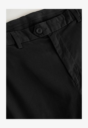 Primo piano della cintura di pantaloni neri con chiusura a bottone, passanti per cintura e cuciture visibili su tessuto liscio.