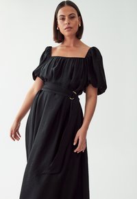 Black maxi dress with a square neckline, puff sleeves, and a belted waist featuring a gold-colored hardware accent. Textured fabric.
