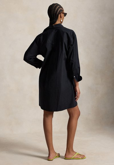 Kvinne med flettehår som har på seg en svart skjortekjole med lange ermer, svarte solbriller, hvite øreringer og gule sandaler, stående med én hånd på hoften.