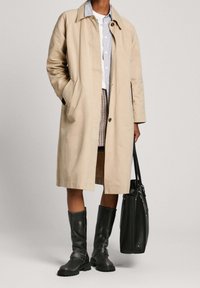 Béžový trenčkot s límcem, bočními kapsami a volným střihem. Nosený přes světle modrou košili a kostkovanou sukni, doplněný černými kozačkami do kolen.