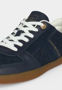 Κοντινό πλάνο σε ναυτικό μπλε sneaker Tommy Hilfiger με λευκά κορδόνια, χρυσά παπούτσια οφθαλμοφορίας, ανάγλυφο λογότυπο στην πλευρική πλευρά και καφέ λαστιχένια σόλα.