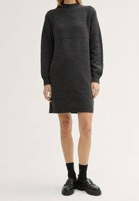 Grijs gebreid jurk met ribbelstructuur, lange mouwen en een ronde hals. Heeft een rechte pasvorm en een zoomlijn op middendij. Gepaard met zwarte schoenen.