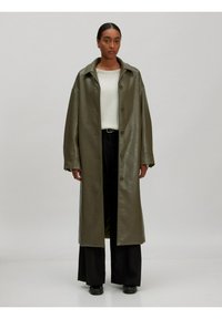 Olijfgroen leren jas met een ontspannen pasvorm, grote kraag en knoopsluiting, gecombineerd met een witte trui en zwarte wijde broek.