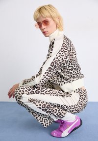 Leopard-print tracksuit with cream accents, fitted design, worn with purple sneakers. Model is crouched against a plain background.