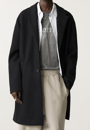 Personne portant un long manteau noir par-dessus un t-shirt graphique gris, une chemise blanche à rayures boutonnée, un pantalon beige, un collier en chaîne argentée et des bagues à la main gauche.