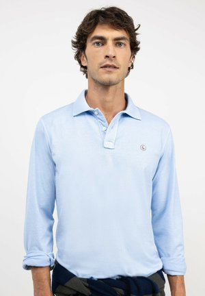 Hombre con cabello rizado oscuro, con una camisa polo azul claro de manga larga y un suéter oscuro atado alrededor de su cintura sobre un fondo blanco.