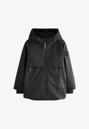 Junge steht und trägt eine schwarze Kapuzenjacke, beige Cargohosen und weiße Sneakers auf einem grauen Boden mit einfarbig grauem Hintergrund.
