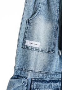 Pantalones de mezclilla azul claro con tirantes ajustables, un bolsillo frontal y una etiqueta de tela con texto, mostrando una textura ligeramente desgastada.