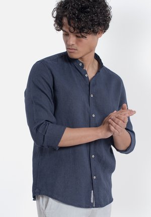 Giovane uomo con capelli ricci che indossa una camicia blu navy con bottoni e maniche arrotolate, guarda verso il basso e si sistema il polso destro.