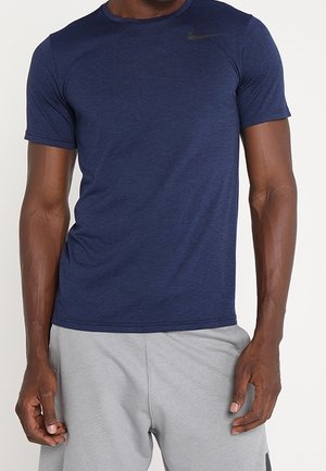 Mężczyzna w granatowej koszulce Nike i jasnoszarych sportowych szortach, stojący z ramionami swobodnie opuszczonymi wzdłuż ciała.