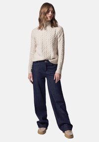 Kremowy sweter w splot warkoczowy z wysokim kołnierzem i długimi rękawami, zestawiony z ciemnoniebieskimi spodniami typu wide-leg i beżowymi butami w stylu mokasynów.
